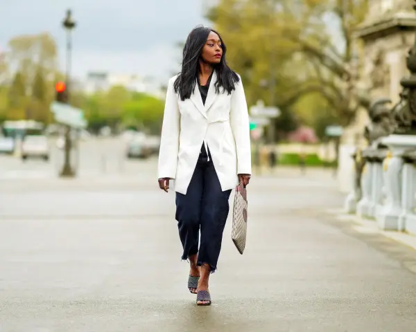 Mujer con estilo **smart casual** camina por la calle con un blazer blanco, jeans oscuros y sandalias de tacón.