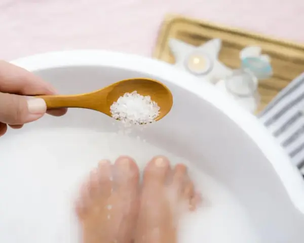 A hand holds a wooden spoon with coarse salt, about to add it to a white basin where feet are soaking. This is a relaxing foot soak with baking soda.