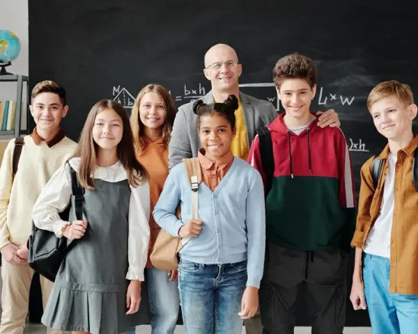 Uśmiechnięty nauczyciel i uczniowie przed tablicą. Czy szkoła społeczna to miejsce, gdzie tak dobrze się czują?