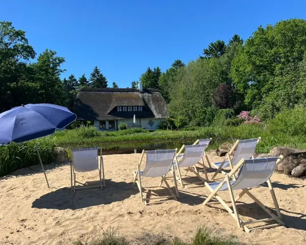 Sonnige Tage am Stedener See: Liegestühle und Sonnenschirm warten auf Besucher vor einem Reetdachhaus.