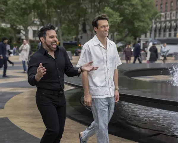 Il cast di And Just Like That, Mario Cantone e Billy Eichner passeggiano ridendo vicino a una fontana in una giornata nuvolosa.