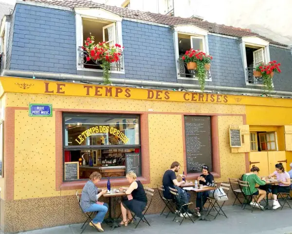 Café "Le Temps des Cerises" con un encantador estilo francés, balcones floridos y gente disfrutando en sus mesas exteriores.