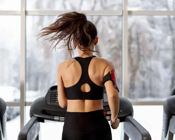 Une femme fait du sport à jeun sur un tapis roulant, ses cheveux flottant au vent. Dehors, la neige tombe.