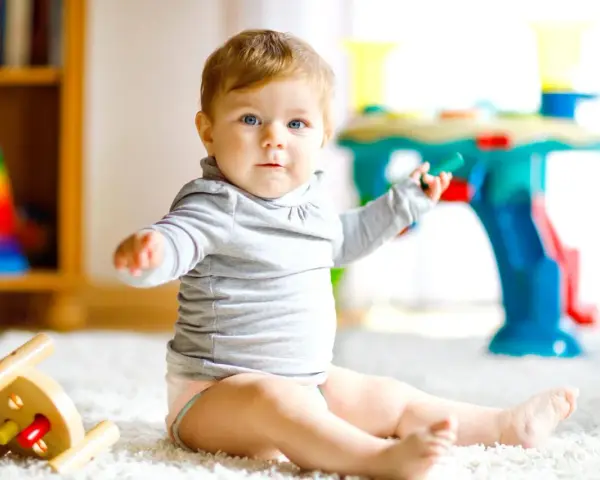 Ein neugieriges 8 Monate altes Baby mit blauen Augen sitzt auf einem flauschigen Teppich und spielt mit Spielzeug.