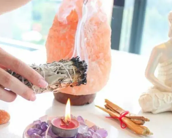 Manos sostienen un manojo de salvia humeante, junto a una lámpara de sal rosa, una vela encendida y una estatua de Buda. Esencial para la limpieza energética de casa.