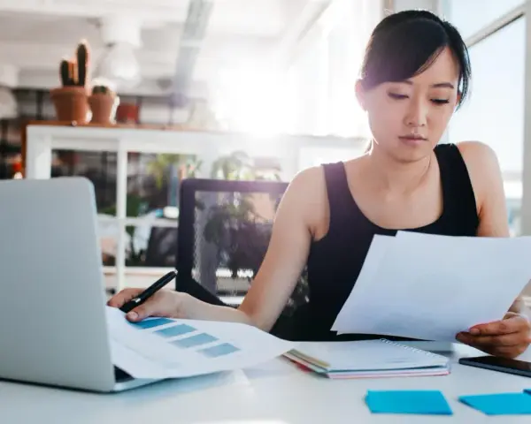 Junge Frau prüft Dokumente, vielleicht die Urlaubsbescheinigung des letzten Arbeitgebers, am Schreibtisch mit Laptop.