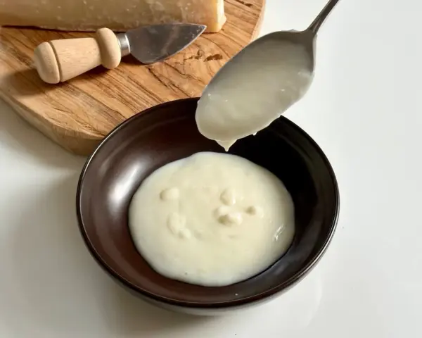 Una cucchiaiata di cremosa crema di parmigiano cade in una ciotola scura. Sul tagliere, un pezzo di parmigiano e un coltellino.