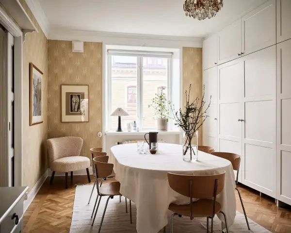 Comedor con mesa cubierta, sillas de madera y pared con papel tapiz dorado. Armarios blancos y suelo de parqué.