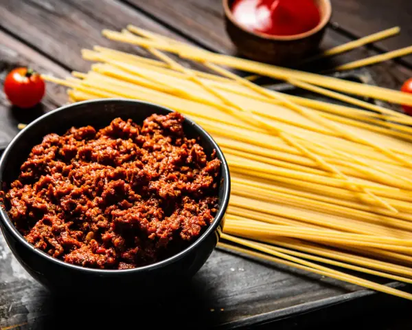 Un tazón de carne para espaguetis a la boloñesa, junto a pasta cruda, tomates y salsa.