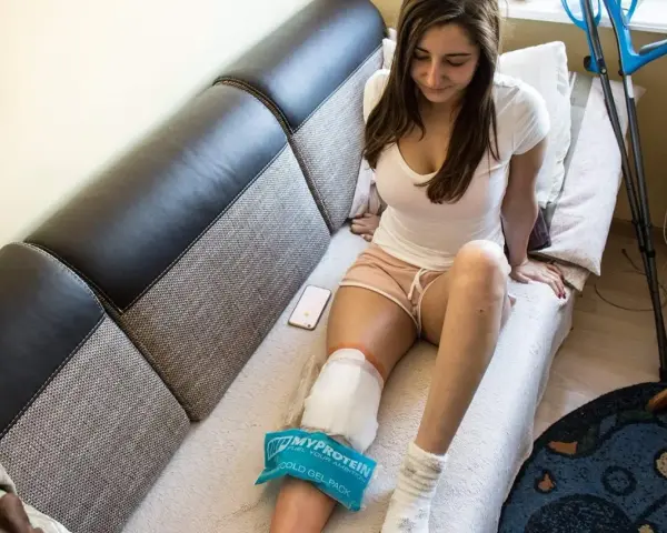 Młoda kobieta z zimnym okładem na kolanie po artroskopii, siedząca na kanapie obok telefonu i kul.