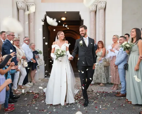 Photographie de mariage : le couple sort de l'église sous une pluie de pétales, entouré de leurs invités joyeux.