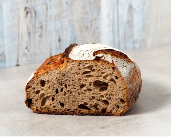 Ein halbes, knuspriges schnelles Sauerteigbrot mit großer Krume und rustikaler Kruste liegt auf einem hellen Untergrund vor einer hellblauen Holzvertäfelung.