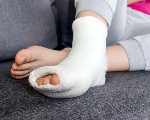 Dziecko z unieruchomioną stopą w białej longecie gipsowej na nodze, odpoczywa na kanapie.
