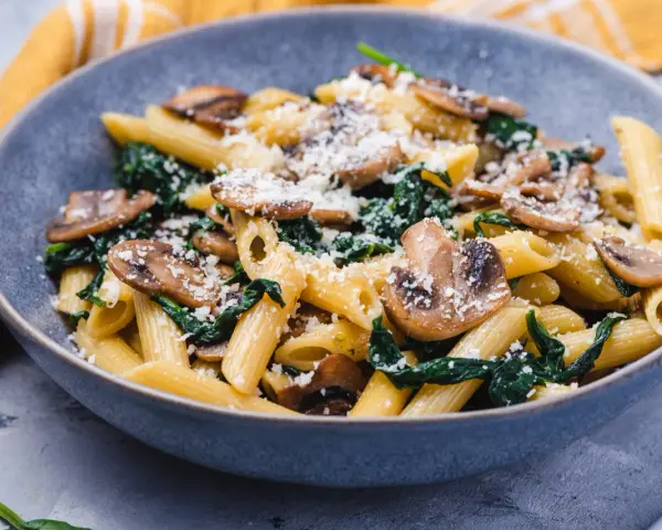 Penne z pieczarkami i szpinakiem, posypane parmezanem, w szarej misce.