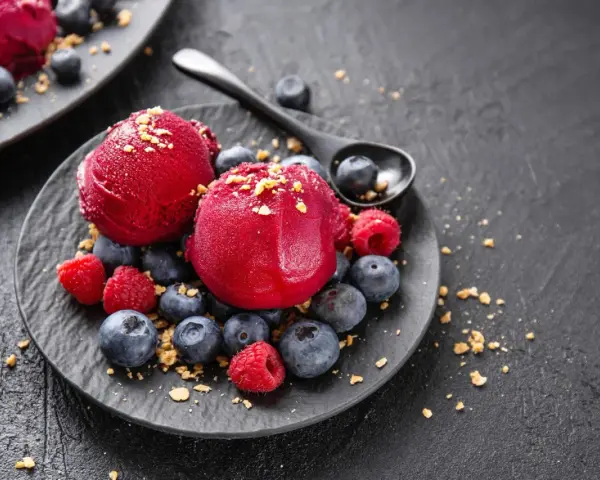 Zwei Kugeln Campari Sorbet mit frischen Beeren und Streuseln auf einem schwarzen Teller.
