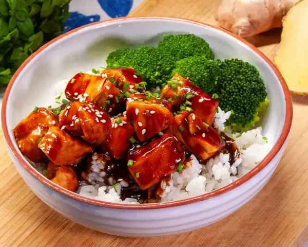 Miseczka ryżu z kawałkami lśniącego lososia teriyaki, posypanego sezamem i szczypiorkiem, obok parujących różyczek brokułów.