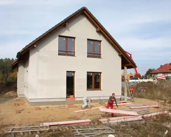 Budowa domu w toku. Pracownicy układają płyty, a w tle widać betonowóz. Czas budowy zależy od wielu czynników.