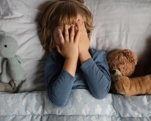 Kind versteckt Gesicht, umgeben von Kuscheltieren. Die Angst um das Kind ist spürbar, es sucht Trost bei seinen Freunden.