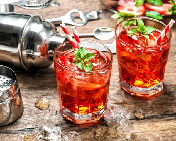 Zwei erfrischende Erdbeer-Cocktails mit Minze und Eiswürfeln, bereit zum Genießen. Ein Shaker und Bar-Werkzeuge deuten auf das kunstvolle Alkohol mischen hin.