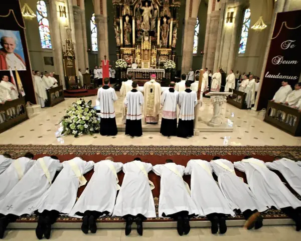 Klerycy w białych szatach leżą na posadzce kościoła, w geście pokory. Obok nich stoją inni duchowni, a w tle widać ołtarz i portret papieża.