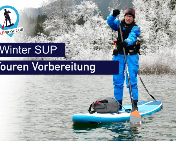Person im blauen Neoprenanzug bereitet sich auf eine Winter SUP Tour vor. Die Landschaft ist mit Schnee bedeckt.