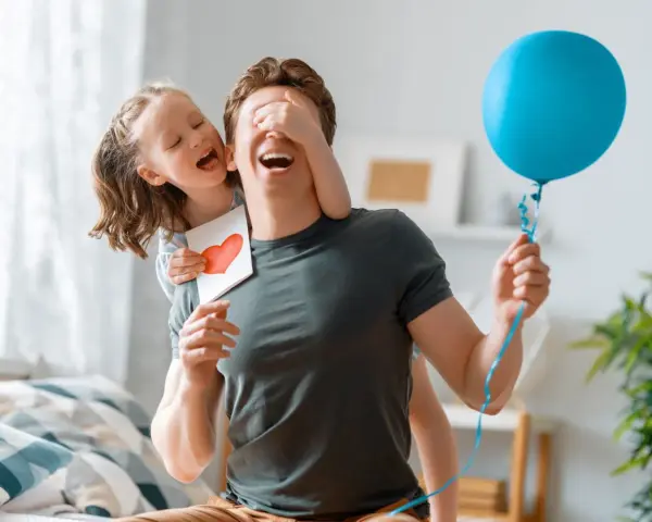 Mała dziewczynka z kartką i sercem zakrywa oczy wujkowi, który trzyma niebieski balon. To piękne życzenia urodzinowe dla wujka!