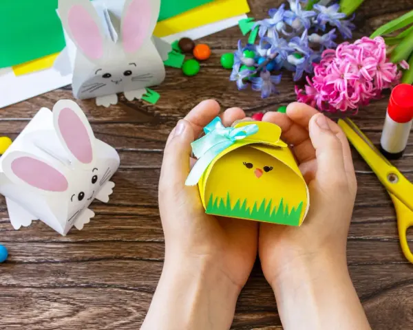Ręce trzymają żółte pudełko w kształcie kurczaka, gotowe jako prezenty na Wielkanoc dla dzieci w szkole. Obok leżą papierowe zajączki i kolorowe cukierki.