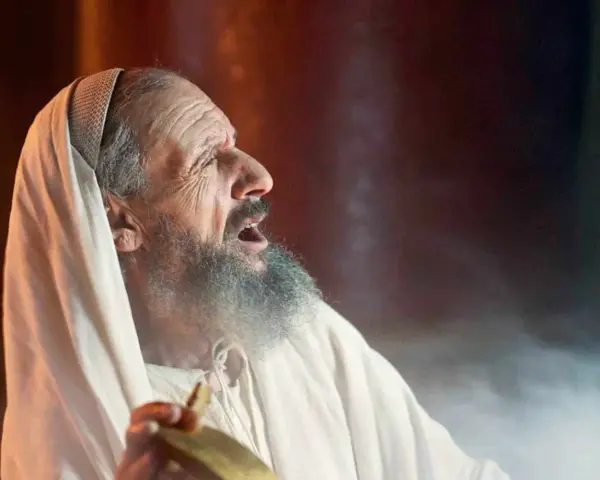 An elderly man, possibly Zechariah in the Bible, with a grey beard and wearing a white robe and head covering, cries out with his mouth open.