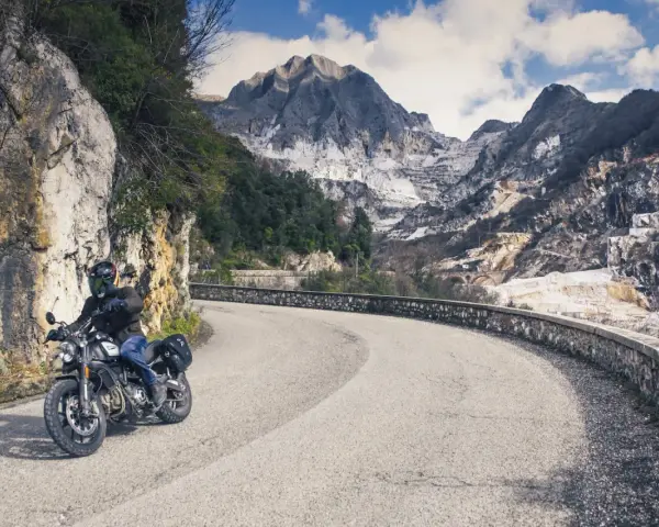 Un motociclista percorre una strada tortuosa tra le montagne della Toscana, con cave di marmo sullo sfondo.