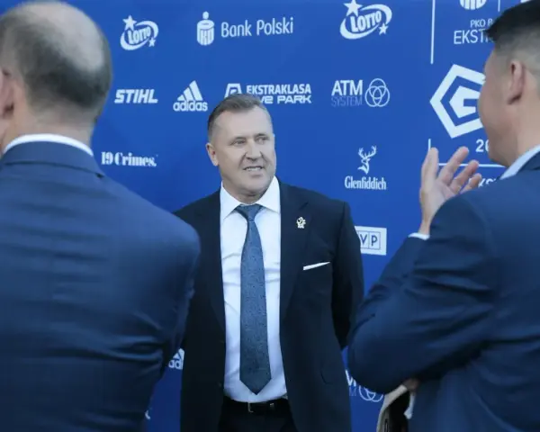 Mężczyzna w garniturze uśmiecha się, stojąc na tle niebieskiej ściany z logotypami sponsorów, być może rozmawiając o tym, ile dostają kluby ekstraklasy.