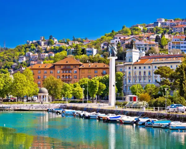 Malowniczy port w Chorwacji z łodziami przycumowanymi do nabrzeża. W tle widać wzgórze z zabudowaniami i zamek.