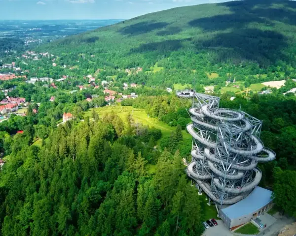 Widok na wieżę widokową w otoczeniu zielonych gór w Polsce. Spiralne schody prowadzą na szczyt, skąd rozpościera się panorama na okoliczne lasy i miasteczko.