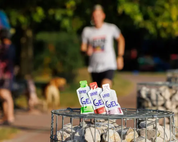 Trzy opakowania żeli energetycznych dla biegaczy: zielone jabłko, cola i truskawka z bananem, na tle biegnącej osoby.