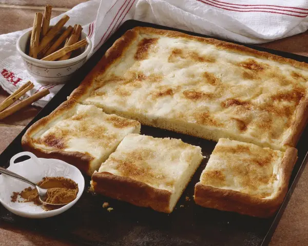 Ein köstlicher Rahmkuchen vom Blech, bestreut mit Zimt und Zucker. Daneben eine Schale mit Zimtstangen und Zimtpulver.