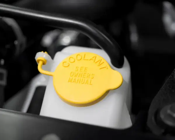 Close-up of a car's coolant reservoir with a yellow cap. The cap reads "COOLANT SEE OWNERS MANUAL," indicating where to find instructions on how to put coolant in car.