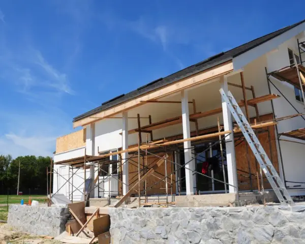 Une maison en construction, avec des échafaudages et une échelle, en cours d'agrandissement. Le ciel bleu est parsemé de nuages légers.
