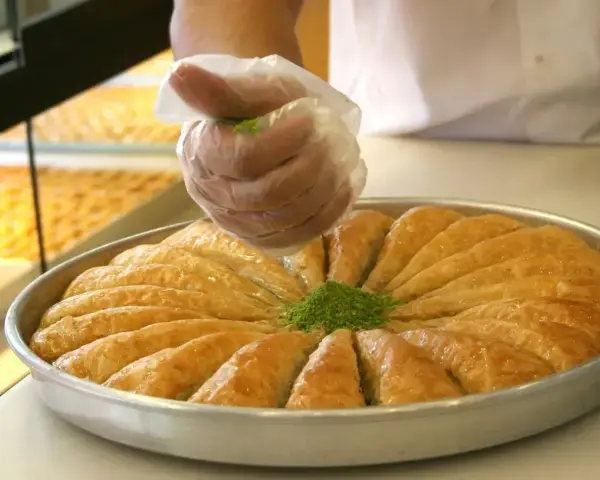 Ręka w rękawiczce posypuje zielonymi pistacjami okrągłą blachę z baklavą. To właśnie baklava co to za deser!