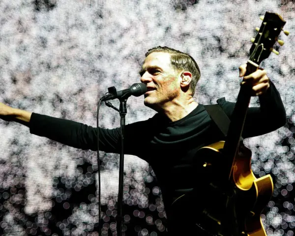 Bryan Adams na koncercie, śpiewa do mikrofonu i gra na gitarze. Wskazuje palcem na publiczność, tworząc niezapomniane chwile.