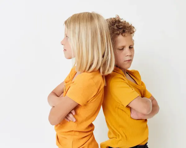 Zwei Kinder stehen mit verschränkten Armen Rücken an Rücken. Ihre Mimik deutet auf **Gewalt unter Geschwistern** hin, ein Streit, der sie trennt.