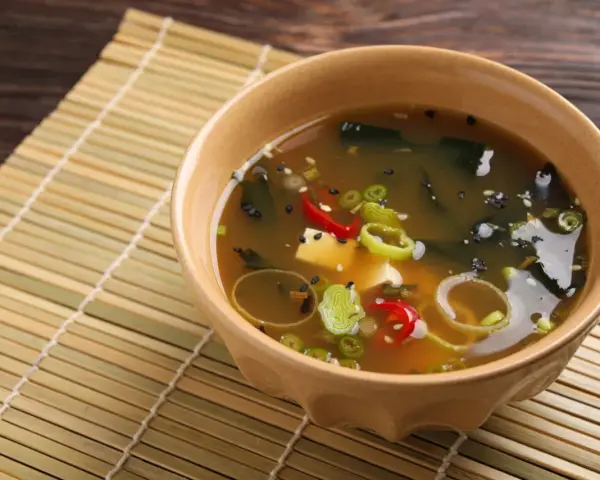 Aksamitna zupa miso z tofu, wodorostami, szczypiorkiem i papryczką chili, podana w ceramicznej miseczce na bambusowej macie.