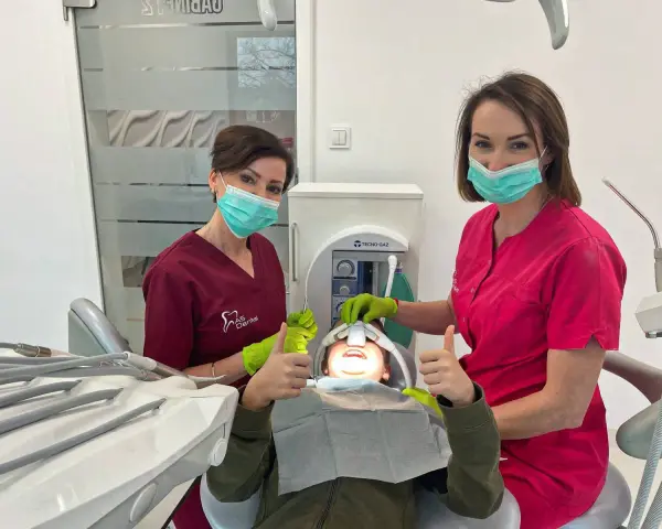 Dwie dentystki w maseczkach i rękawiczkach z uśmiechniętym pacjentem w gabinecie stomatologicznym.