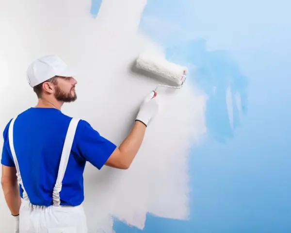 Un peintre en bleu et blanc applique une nouvelle couche de peinture bleue sur un mur blanc. C'est une peinture sur peinture en cours.