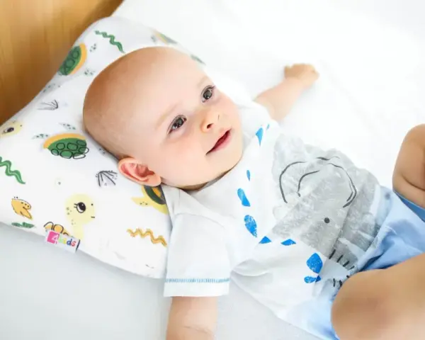 Uśmiechnięty bobas leży na białej pościeli, opierając główkę na poduszce ortopedycznej dla niemowląt z motywem żółwi.