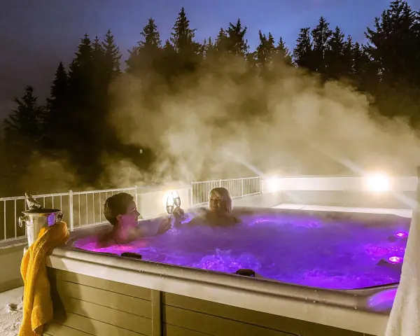 Zwei Frauen stoßen mit Champagner in einem beleuchteten Whirlpool an, umgeben von Dampf und Bäumen. Ein perfektes Wochenende mit Gott.