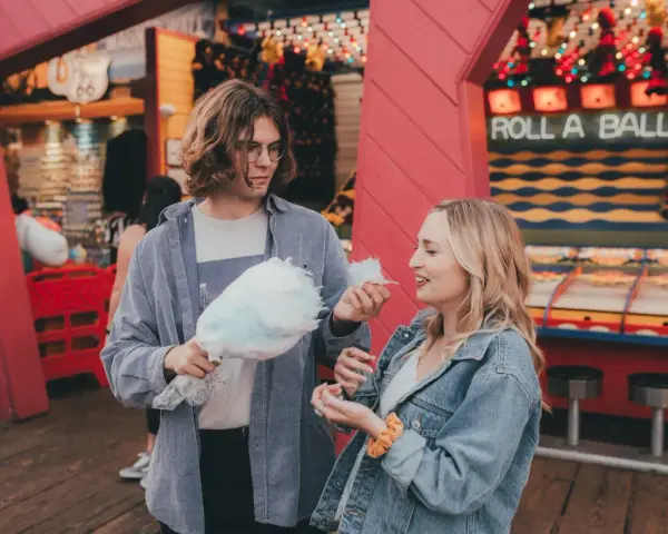 Ein Paar teilt sich Zuckerwatte vor einem "Roll a Ball"-Spiel im Freizeitpark. Ihr lässiger Freizeitpark-Outfit passt perfekt zum Tag.