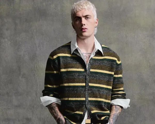 Joven con pelo rubio platino y tatuajes lleva un jersey de rayas verdes y amarillas, mostrando que **que jerseys se llevan este año** son los de estilo retro.
