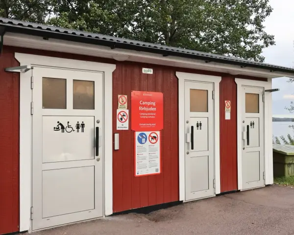 Röd byggnad med toaletter vid en sjö. Skyltar visar att camping är förbjuden. En trevlig rastplats med toalett för resenärer.