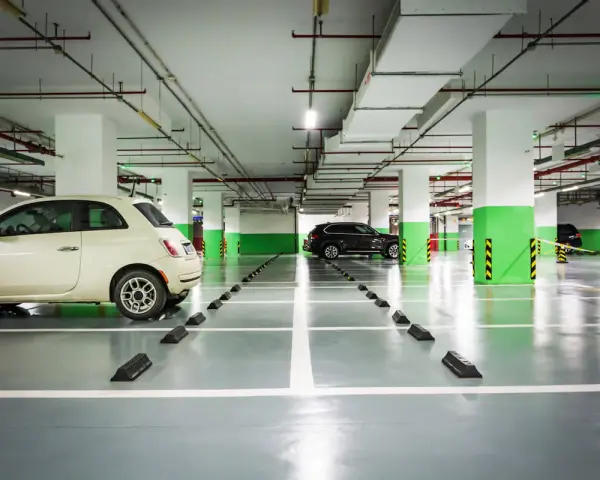 Jasny samochód i ciemny SUV stoją na parkingu podziemnym. Widać wyraźnie wyznaczoną szerokość miejsca parkingowego.