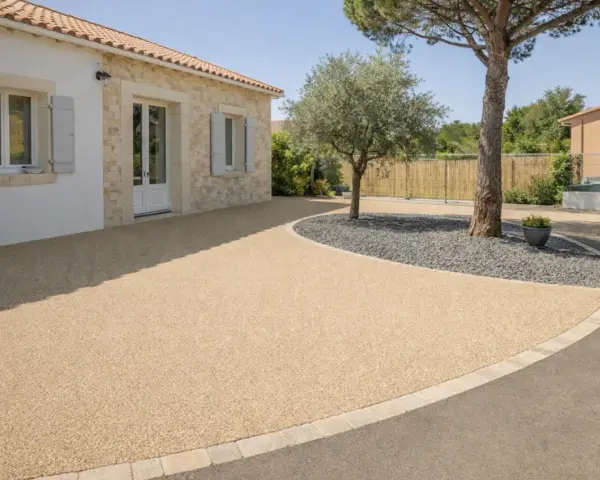 Allée de maison avec béton désactivé de belle épaisseur, bordée de pierres et de gravier sombre autour d'un arbre.