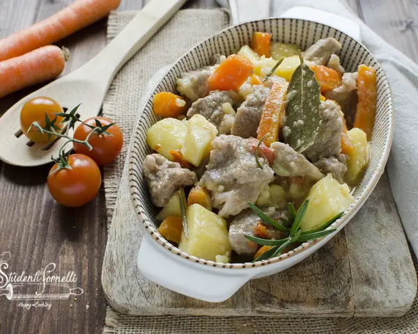 Un invitante spezzatino in bianco con patate e carote, guarnito con rosmarino e alloro. Accanto, pomodorini e carote fresche.
